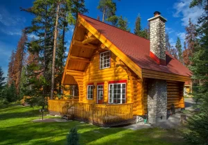 Exterior of cabin at Alpine Village Jasper. The cabin features a large wrap-around wooden deck and is surrounded by lush green grass and tall trees under a clear blue sky.