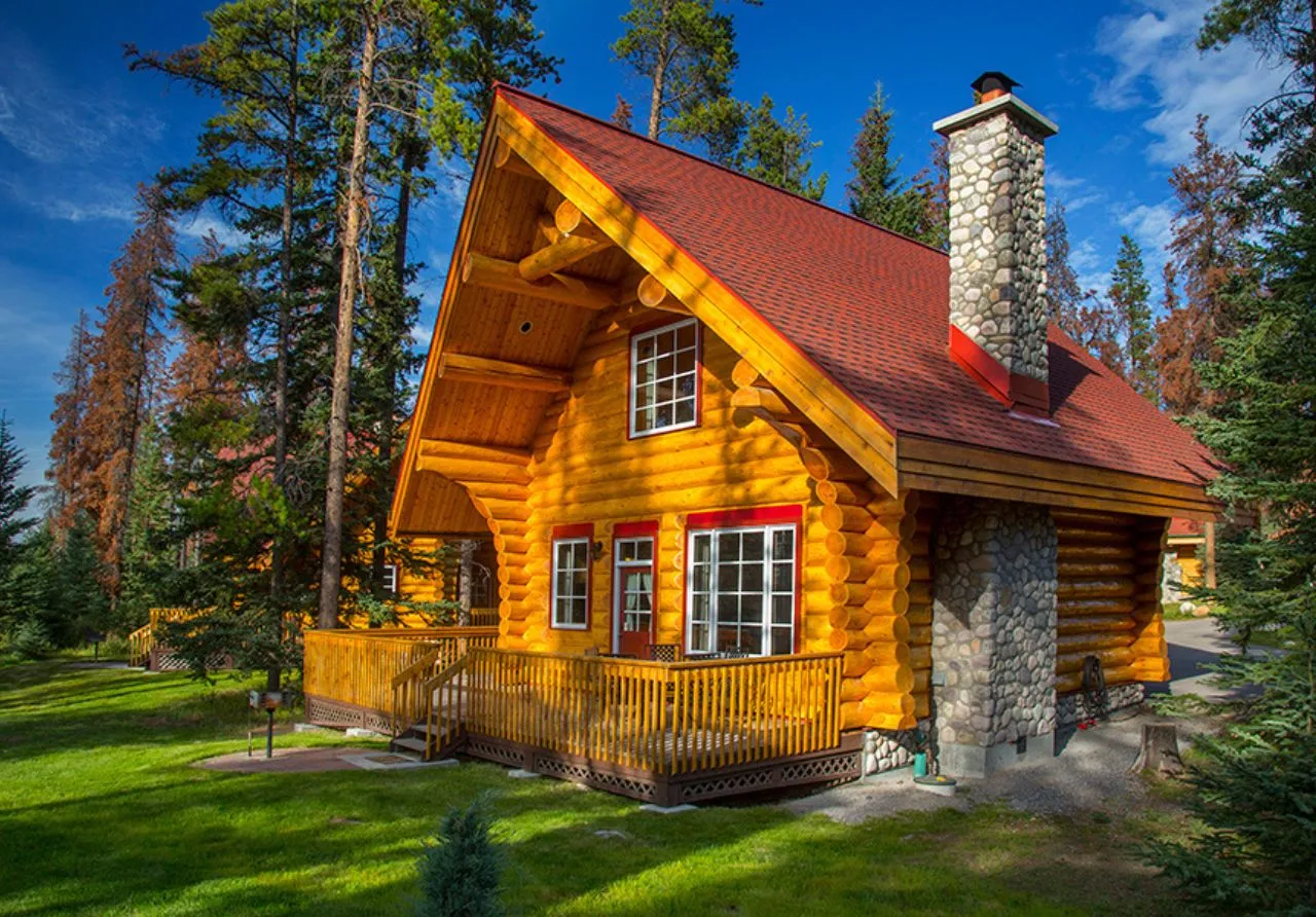 Exterior of cabin at Alpine Village Jasper. The cabin features a large wrap-around wooden deck and is surrounded by lush green grass and tall trees under a clear blue sky.