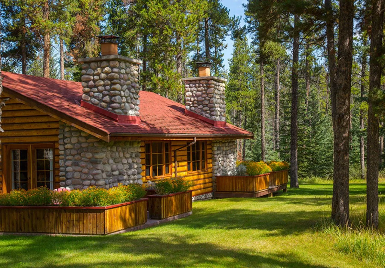 A long, rustic cabin at Alpine Village featuring two large, prominent stone chimneys and a red roof. Bright colour flowers fill large wooden planter boxes that line the base of the cabin. The building is surrounded by green grass and sits at the edge of a dense forest.