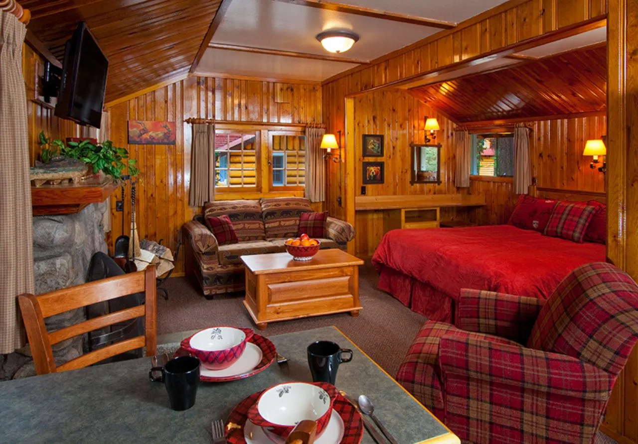 Interior of a Heritage One Room Cabin at Alpine Village, featuring warm wood-paneled walls and ceilings. The open-concept space in this image includes a stone fireplace with a mounted TV, a patterned sofa, a bed with a red quilt, and a red plaid armchair. In the foreground, a dining table is set with plaid-patterned bowls and black mugs.