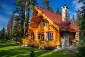 Exterior of cabin at Alpine Village Jasper. The cabin features a large wrap-around wooden deck and is surrounded by lush green grass and tall trees under a clear blue sky.