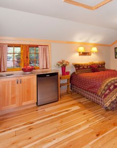 Interior of a cozy one-room cabin featuring a bed with a rustic moose-patterned bedspread, hardwood floors, and a small refrigerator