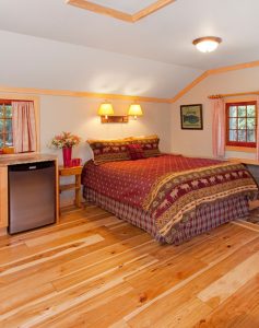 Interior of a cozy one-room cabin featuring a bed with a rustic moose-patterned bedspread, hardwood floors, and a small refrigerator
