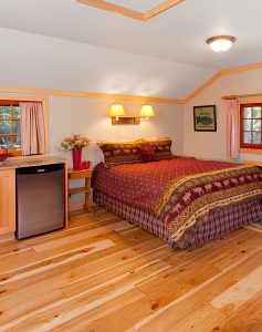 Interior of a cozy one-room cabin featuring a bed with a rustic moose-patterned bedspread, hardwood floors, and a small refrigerator